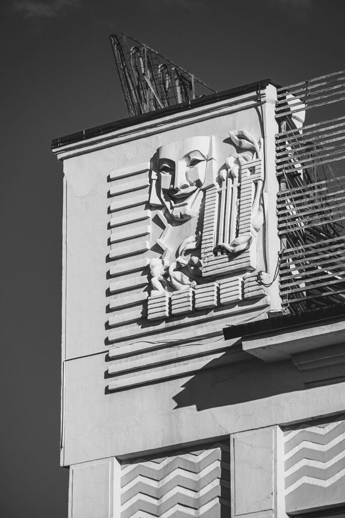 Black and white image showing the front of a theatre with a masoned happy face and other decoration on it